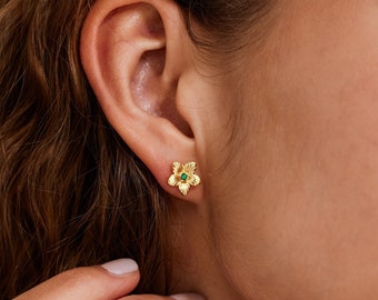 May include: A gold stud earring shaped like a five-petaled flower with a small, round, green gemstone set in the center. The metal surface of the flower petals features fine, radiating textured lines that extend from the center stone outward.