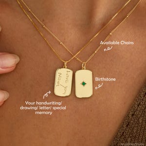 Op de afbeelding: Twee goudkleurige kettingen met rechthoekige hangers. Eén hanger is voorzien van een gegraveerd cursief handschrift met de tekst 'love you Nana', terwijl de andere een kleine, centraal geplaatste groene edelsteen in een stervormig metalen detail bevat.