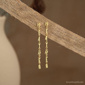May include: Gold-colored dangle earrings spelling out the name "HEATHER" in capital letters. The earrings are suspended from a delicate chain and are displayed against a neutral background.