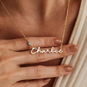 Peut inclure: Collier en or avec un pendentif en forme de plaque gravée du nom "Charlie" en cursive et une petite pierre bleue.