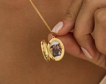 May include: A gold-toned oval locket pendant suspended on a delicate matching chain. The locket is open, revealing a small, rectangular black and white photograph of two individuals inside the polished metal frame. The piece features a classic hinge.