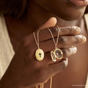 May include: Two gold oval lockets on delicate chains. One locket is closed and features a black cross symbol on the front. The second locket is open, revealing two small oval photographs inside, showing portraits of people within the gold-toned frame.