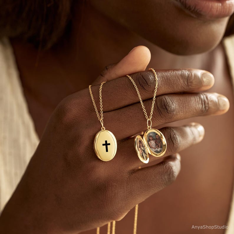 May include: Two gold oval lockets on delicate chains. One locket is closed and features a black cross symbol on the front. The second locket is open, revealing two small oval photographs inside, showing portraits of people within the gold-toned frame.