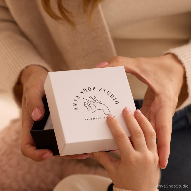 May include: A white square gift box with a black interior is being opened. The box has a logo that says "ANYA SHOP STUDIO HANDMADE JEWELRY" with a hand holding a crystal. The box is being held by two people.