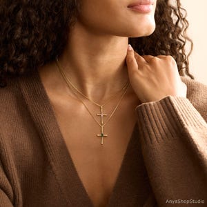 May include: Two gold-colored cross necklaces. One cross features a pink gemstone, and the other has a green gemstone. The necklaces are layered and worn on a person with brown hair and a brown sweater.