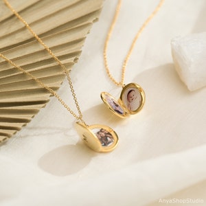 May include: Two gold-toned oval locket necklaces with delicate chains. One locket is open, revealing a photograph of a baby, while the other is partially open, showing a photograph of two people and the letter M engraved on the exterior surface.