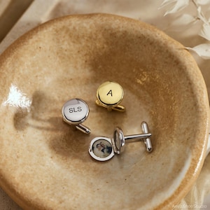 May include: Three round cufflinks: one silver-toned engraved with SLS, one gold-toned engraved with the letter A, and one silver-toned locket-style cufflink containing a small circular photograph of two people inside the open casing.