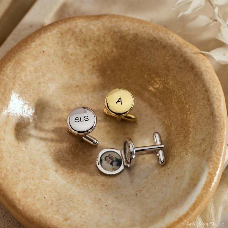 May include: Three round cufflinks: one silver-toned engraved with SLS, one gold-toned engraved with the letter A, and one silver-toned locket-style cufflink containing a small circular photograph of two people inside the open casing.