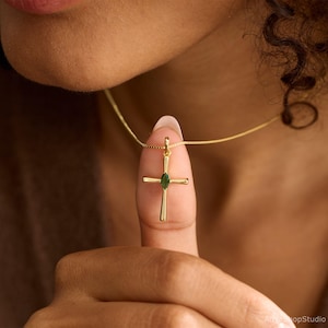 May include: A gold-colored cross pendant necklace with a small, oval-shaped green gemstone at the center. The cross is suspended from a delicate gold chain. The image is a close-up, highlighting the jewelry's details.