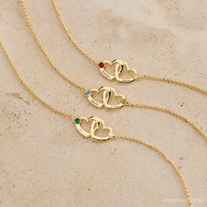 May include: Three gold-tone necklaces, each featuring two interlocking heart charms. Each heart is engraved with a name and accented with a small round gemstone. The necklaces have delicate gold-tone chains and are displayed in a parallel arrangement.