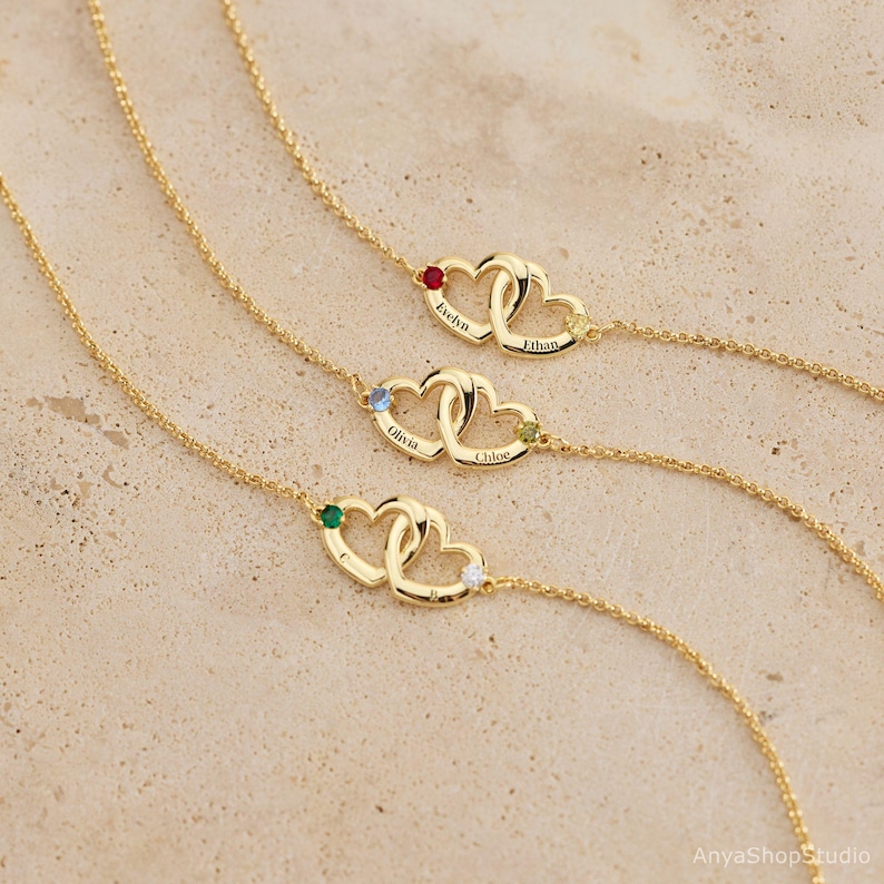 May include: Three gold-tone necklaces, each featuring two interlocking heart charms. Each heart is engraved with a name and accented with a small round gemstone. The necklaces have delicate gold-tone chains and are displayed in a parallel arrangement.