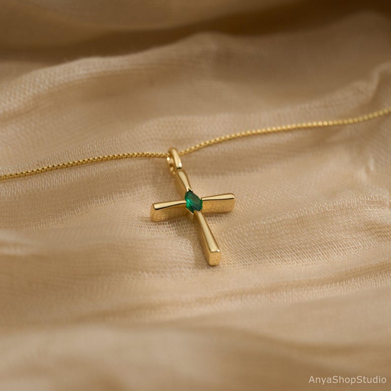 May include: A gold-colored cross pendant necklace with a small, green gemstone at the center. The cross is suspended from a delicate chain. The background is a soft, neutral fabric.