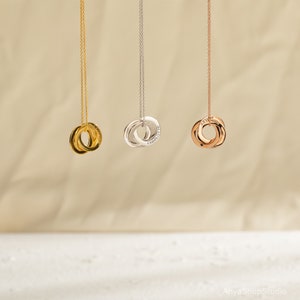 May include: Three gold, silver, and rose gold necklaces with interlocking rings. Each ring is engraved with a name. The necklaces are hanging against a light beige background.