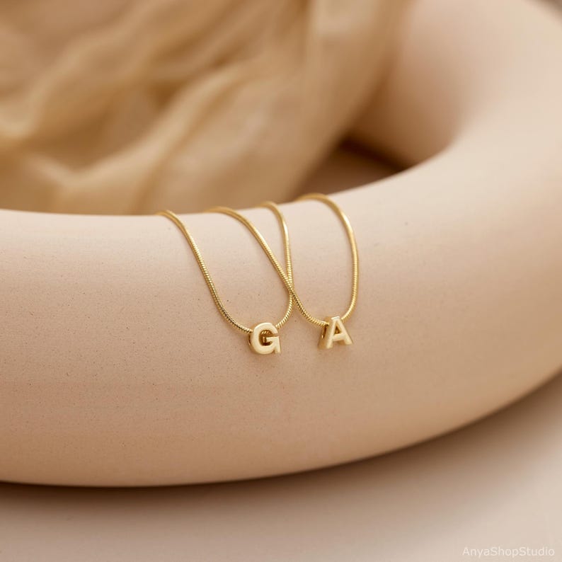 May include: Two gold-toned snake chain necklaces, each featuring a single capital letter pendant. One necklace displays the letter G and the other displays the letter A. The necklaces are draped over a smooth, curved, beige-colored object.