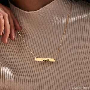 May include: Gold bar necklace with the Arabic word "فاطمة" engraved on it.