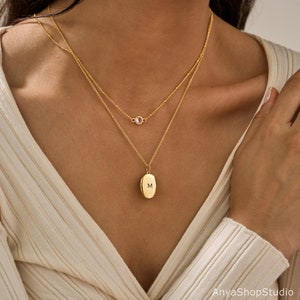 May include: A layered gold necklace set featuring two delicate chains. The shorter chain holds a small, round, light pink gemstone. The longer chain features an oval-shaped gold pendant engraved with the capital letter M in the center.