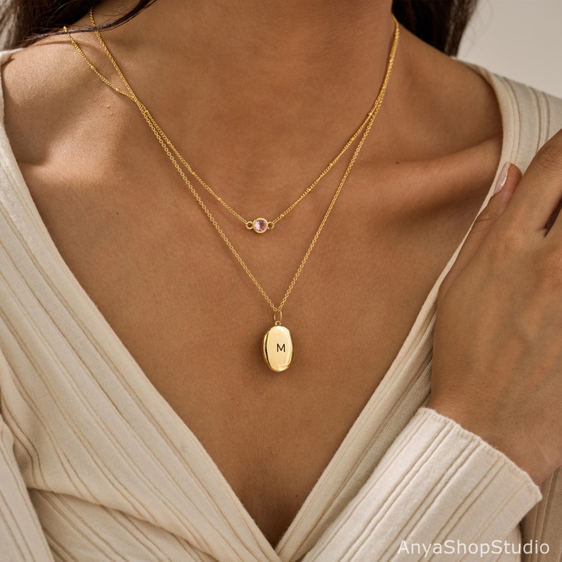 May include: A layered gold necklace set featuring two delicate chains. The shorter chain holds a small, round, light pink gemstone. The longer chain features an oval-shaped gold pendant engraved with the capital letter M in the center.