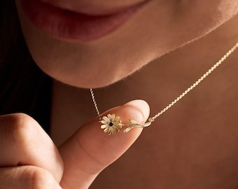 May include: A delicate gold necklace featuring a daisy pendant with a dark center stone. The daisy is attached to a slender chain, creating an elegant and feminine piece of jewelry. The necklace is shown close up, highlighting the intricate details of the floral design.