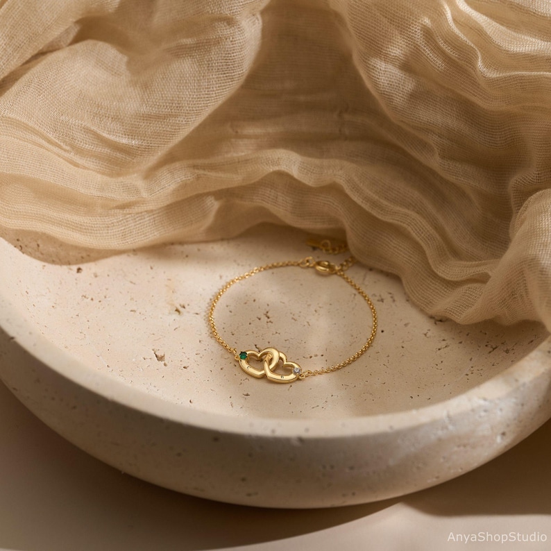 May include: A gold-toned chain bracelet featuring two interlocking heart-shaped charms. One heart is accented with a small green gemstone, while the other heart is accented with a small clear gemstone. The bracelet has a delicate link chain design.