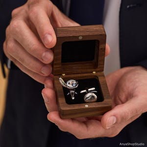 May include: A pair of silver-toned circular cufflinks housed in a dark brown wooden box. One cufflink features an open locket design revealing a small photograph of two people, while the other is a solid silver-toned disc with engraved initials.