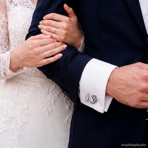 May include: A silver circular cufflink attached to a white shirt cuff, featuring a small, clear-covered locket design that displays a miniature black and white photograph of a couple inside the metal frame.