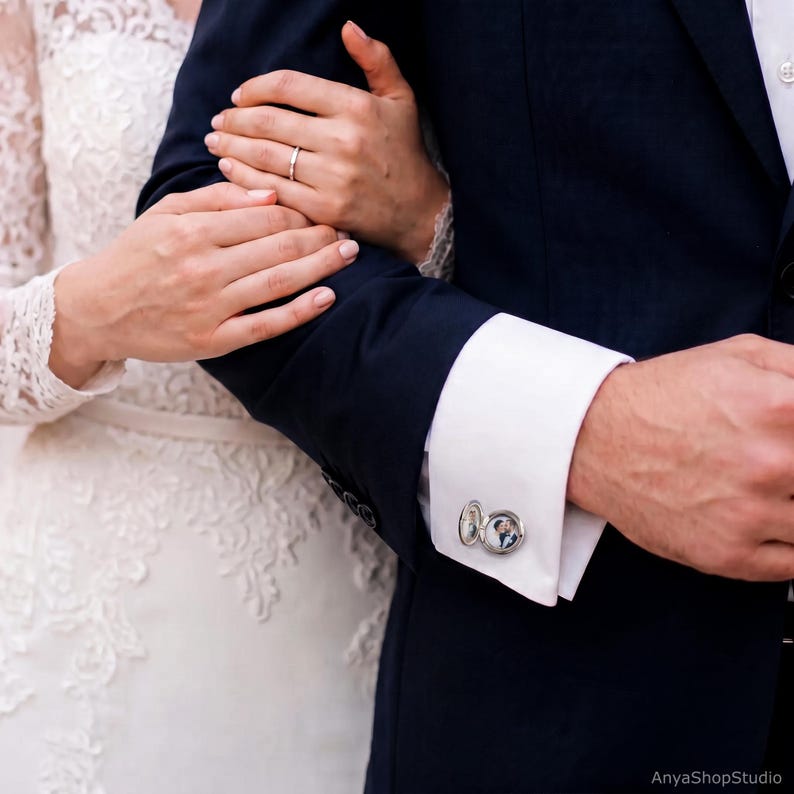 May include: A silver circular cufflink attached to a white shirt cuff, featuring a small, clear-covered locket design that displays a miniature black and white photograph of a couple inside the metal frame.