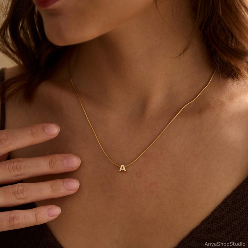 May include: A gold-toned necklace featuring a delicate snake chain and a small, centered letter A pendant. The pendant is polished and minimalist in design, resting against the collarbone. The chain is thin and smooth, providing a subtle shine.