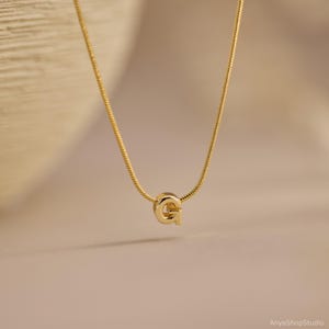 May include: A gold-toned snake chain necklace featuring a single, polished letter G pendant centered on the chain. The metal has a smooth, reflective finish, and the letter is designed in a clean, sans-serif font style, hanging from the thin chain.