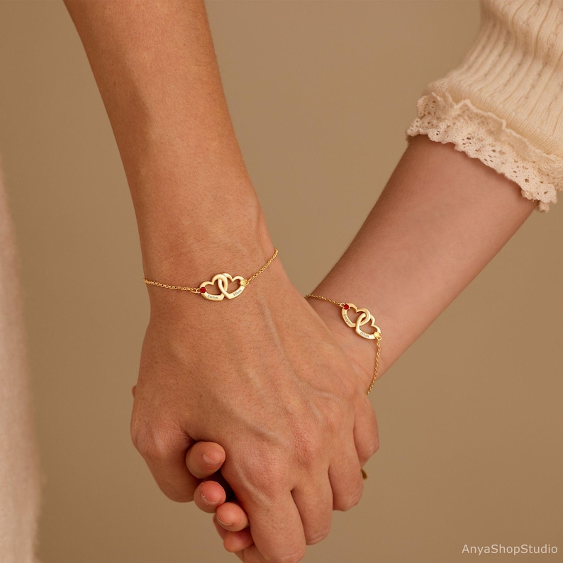 May include: Two gold-toned chain bracelets, each featuring a pair of interlocking heart charms. One heart on each bracelet is accented with a small red stone. The metal hearts are engraved with names, and the bracelets are worn on two wrists.