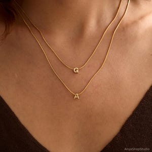 May include: A layered gold-tone necklace set featuring two delicate snake chains. The shorter chain displays a small, polished letter G pendant, while the longer chain features a small, polished letter A pendant, both centered on the chains.