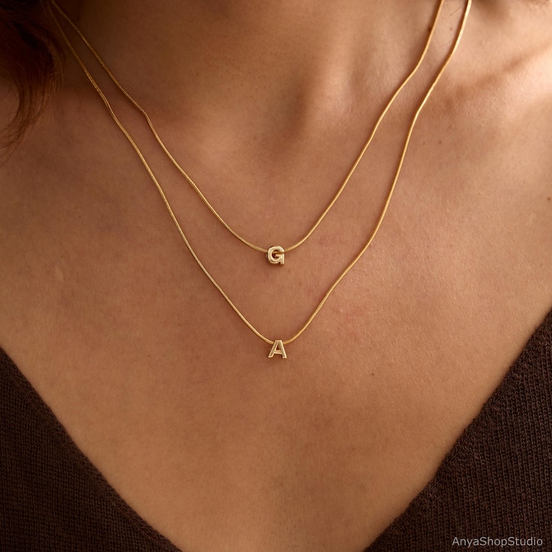 May include: A layered gold-tone necklace set featuring two delicate snake chains. The shorter chain displays a small, polished letter G pendant, while the longer chain features a small, polished letter A pendant, both centered on the chains.