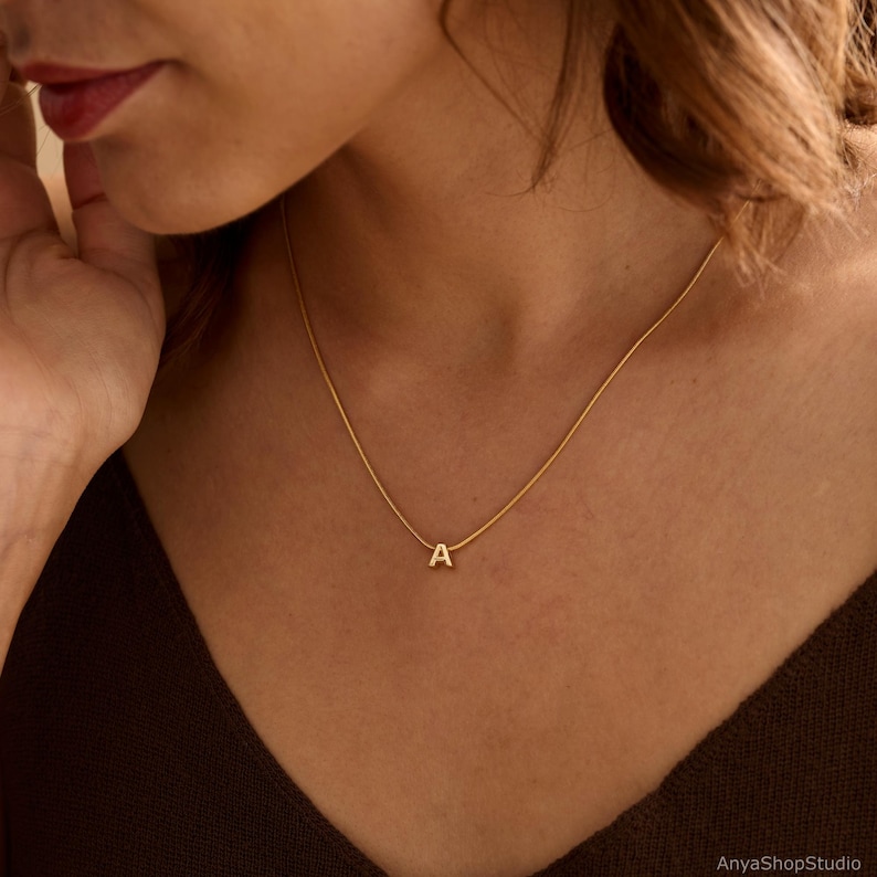 May include: A gold-toned necklace featuring a thin, smooth chain and a small, centered pendant in the shape of the capital letter A. The piece has a polished finish and rests against the collarbone, providing a simple and clean aesthetic.