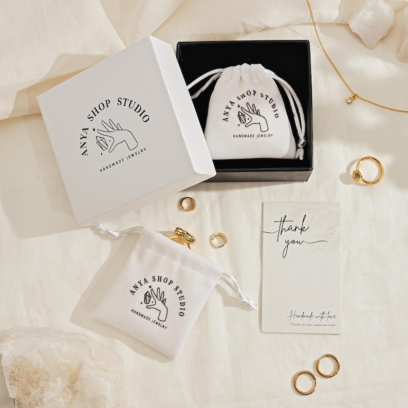 May include: A jewelry set featuring gold rings, a necklace, and small white drawstring bags and a white box with the text "ANYA SHOP STUDIO HANDMADE JEWELRY". A thank you card is also included.