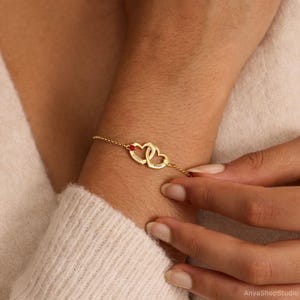 May include: A gold-toned chain bracelet featuring two interlocking heart-shaped charms. Each heart is engraved with a name, and one heart is accented with a small, round, red gemstone. The bracelet is secured with a delicate link chain.