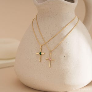 May include: Two gold-colored cross necklaces with delicate chains. One cross features a green gemstone, the other a pink gemstone. The necklaces are displayed against a neutral-toned, textured background, showcasing the jewelry's elegant design.