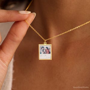 Peut inclure: Un collier doré avec un petit pendentif carré. Le pendentif présente une photographie de deux personnes et le texte manuscrit "Love You" et une date. Le collier est tenu par une personne.