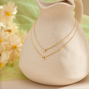 May include: Two gold-toned necklaces featuring thin chains, each adorned with a single letter pendant. The top necklace displays the letter A, while the bottom necklace features the letter G. The jewelry is draped over a cream-colored ceramic pitcher.