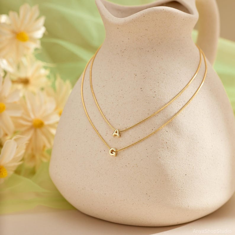 May include: Two gold-toned necklaces featuring thin chains, each adorned with a single letter pendant. The top necklace displays the letter A, while the bottom necklace features the letter G. The jewelry is draped over a cream-colored ceramic pitcher.