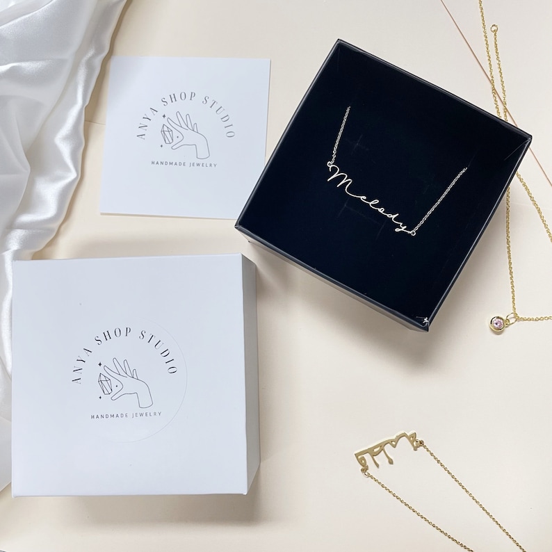 May include: Two white boxes with a black and white logo that says "Anya Shop Studio Handmade Jewelry". A black box with a gold necklace that says "Melody" inside. A gold necklace with a cursive name on it.