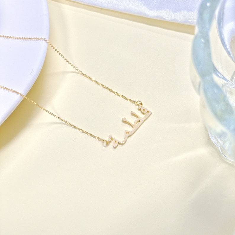 May include: A gold chain necklace with a pendant featuring the Arabic word "Khalid" in a simple, modern font.