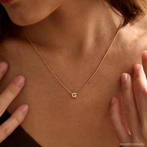 May include: A delicate gold-toned necklace featuring a small, textured letter G pendant suspended from a thin, matching gold-toned chain. The pendant is centered on the chain, which rests against the collarbone area, showcasing a simple design.