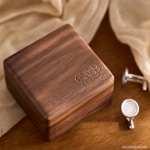 May include: A square dark brown wooden box with a hinged lid featuring the engraved text C&B and the date 27.10.26. Beside the box are two silver-toned metal cufflinks with circular faces, each displaying an engraved name.
