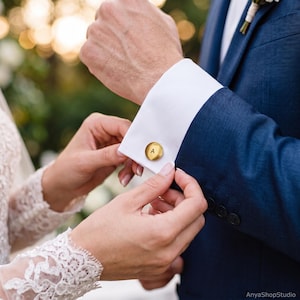 May include: A round, brushed gold-tone metal cufflink featuring the letter A engraved in the center. The cufflink is secured through the white fabric cuff of a navy blue suit jacket sleeve, showcasing a polished and metallic finish.