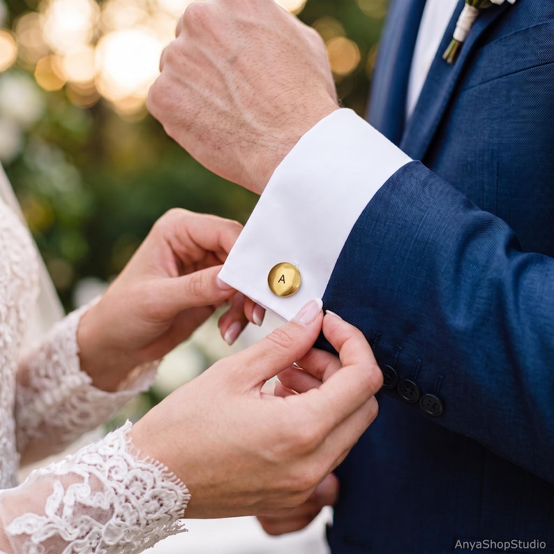 May include: A round, brushed gold-tone metal cufflink featuring the letter A engraved in the center. The cufflink is secured through the white fabric cuff of a navy blue suit jacket sleeve, showcasing a polished and metallic finish.