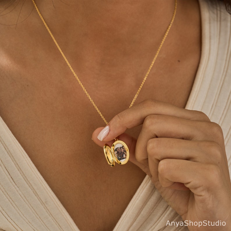 May include: A gold oval locket pendant on a delicate gold chain. The locket is open, revealing a small photograph of two people inside. The metal has a polished, reflective finish. The text AnyaShopStudio is visible in the bottom right corner.
