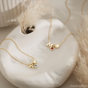 May include: Two gold necklaces with heart charms and initial pendants. The necklace on the left has a blue gemstone and the letter "R". The necklace on the right has a red gemstone and the letter "M".