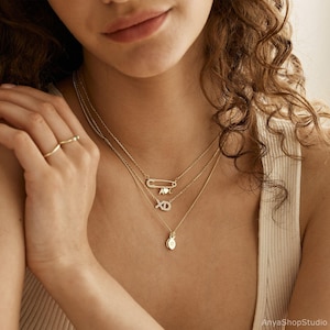 May include: A woman wearing a layered gold necklace with a safety pin charm, a heart charm, and a diamond XO charm. The necklace also features a small round pendant.