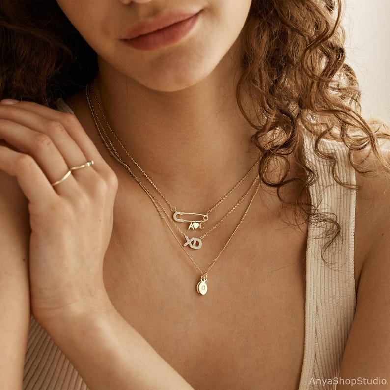 May include: A woman wearing a layered gold necklace with a safety pin charm, a heart charm, and a diamond XO charm. The necklace also features a small round pendant.