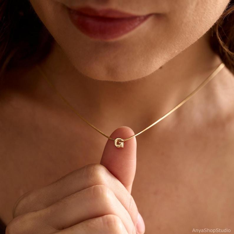 May include: A gold-toned necklace featuring a delicate chain and a small, centred letter G pendant. The pendant is a polished, metallic initial charm suspended on a thin, smooth chain that rests against the neck, showcasing a simple design.