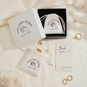 May include: A white jewelry box with a black interior containing a white drawstring pouch, a separate white pouch, several gold rings, a gold necklace with a small pendant, and a thank you card. All items feature the Anya Shop Studio logo.